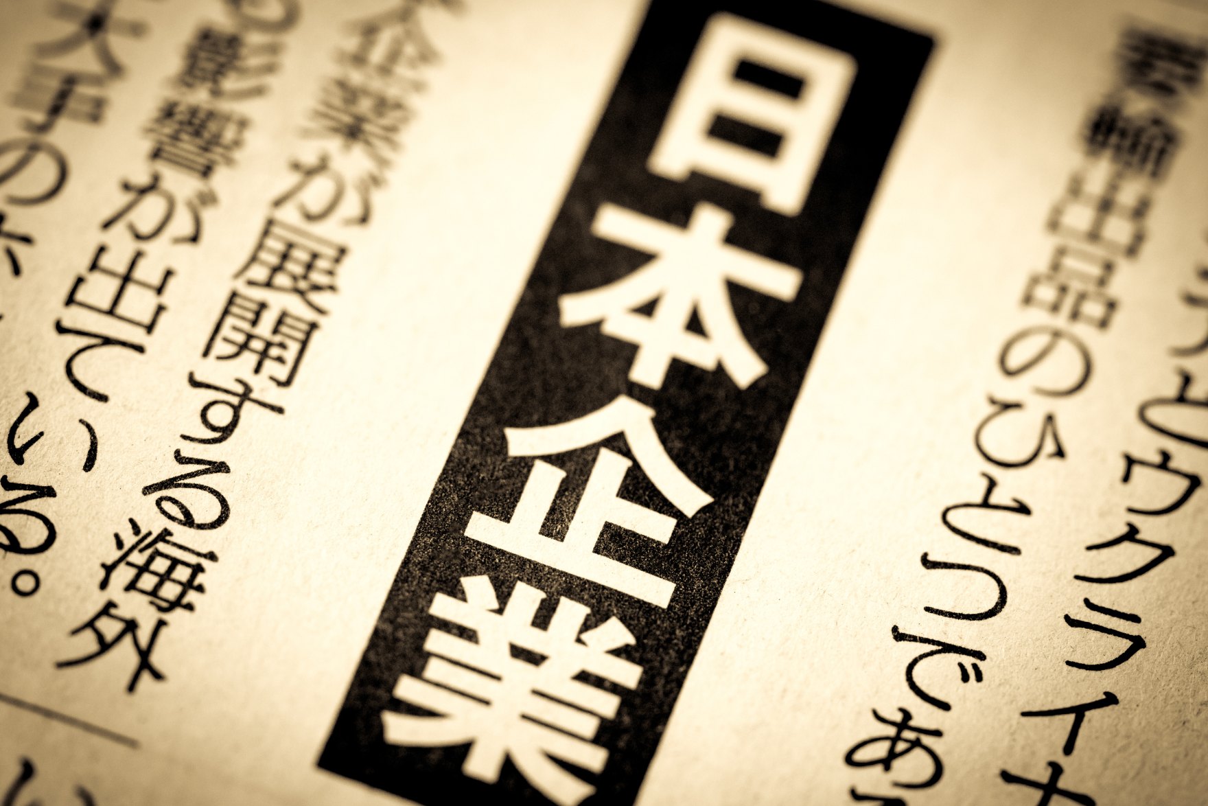 A close-up photo of a Japanese newspaper with the word "日本企業" (Japanese company) in bold, black font. The surrounding text is blurred and unreadable.
