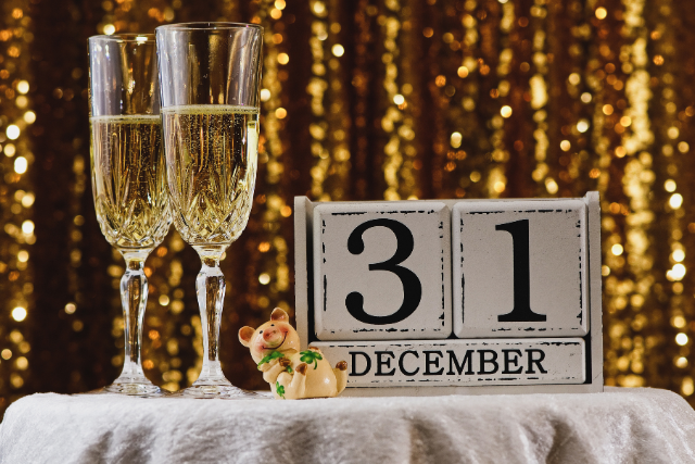 The image depicts a festive scene with two champagne glasses on a table, a calendar showing "December 31," and a plush toy bear. The golden lights in the background add to the celebratory atmosphere, evoking joy and warmth. This image is perfect for promoting a New Year's Eve celebration or conveying a sense of holiday cheer and togetherness.
