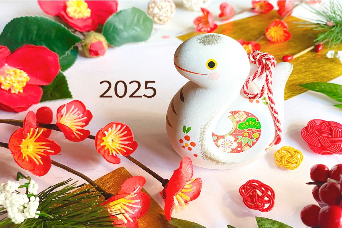 A charming white ceramic nengajo, shaped like a bird and adorned with a red and green decorative pattern. The year "2025" is displayed prominently. Surrounding the nengajo are red flowers and strings of red and white beads, creating a festive and celebratory atmosphere.