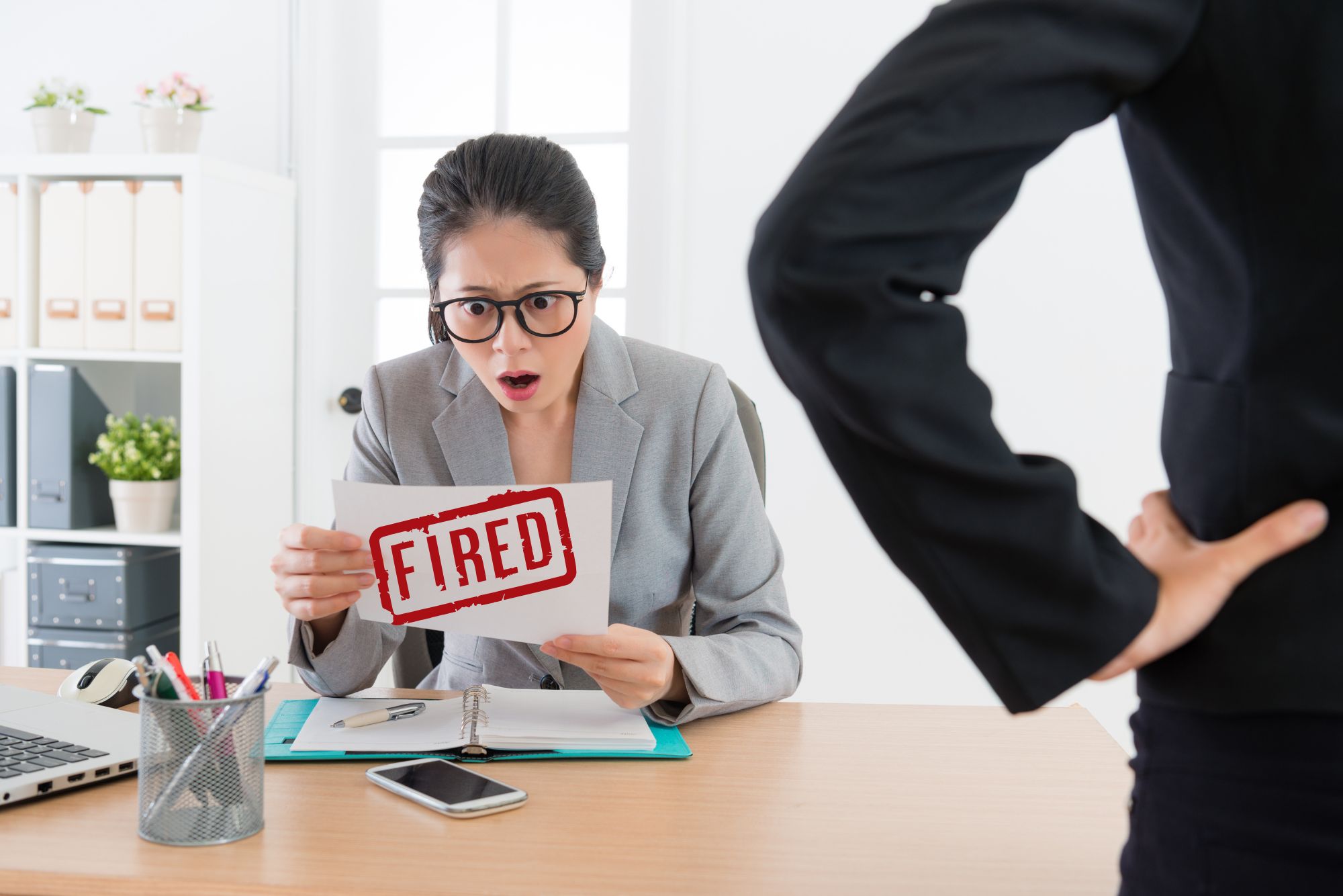 Shocked businesswoman holding a 'FIRED' notice, with a supervisor standing behind her.